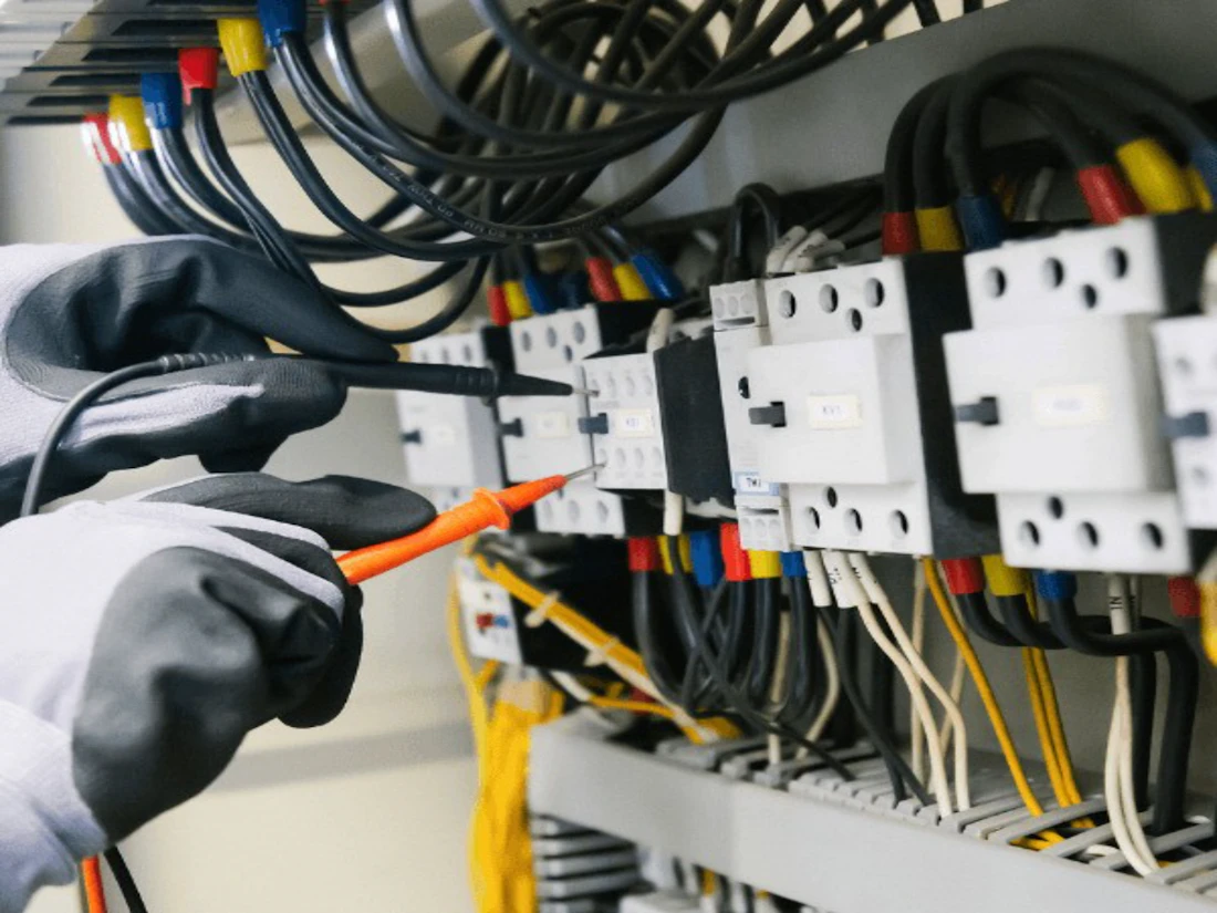Gloved hands testing wiring on an electrical panel with a probe