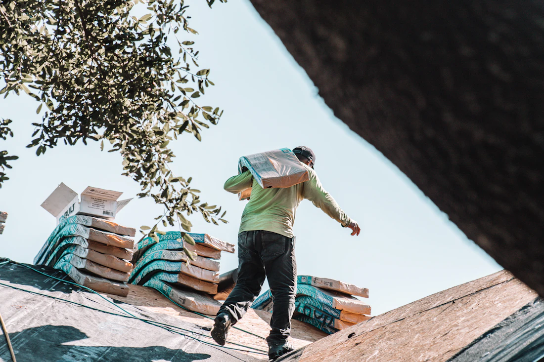 A roofer carrying a bundle of shingles across a residential roof