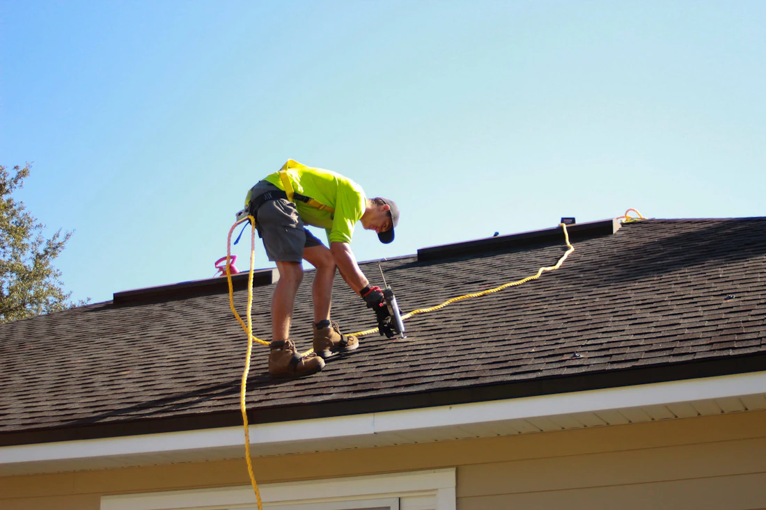 A roofer in a harness working with a power tool on a shingle roof